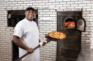 Frank Pepe Pizzeria chef smiles while posing with a pizza next to the coal fire oven